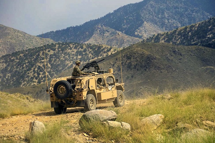 A soldier looks out from a humvee vehicle with green, rolling hills and mountains in the background.