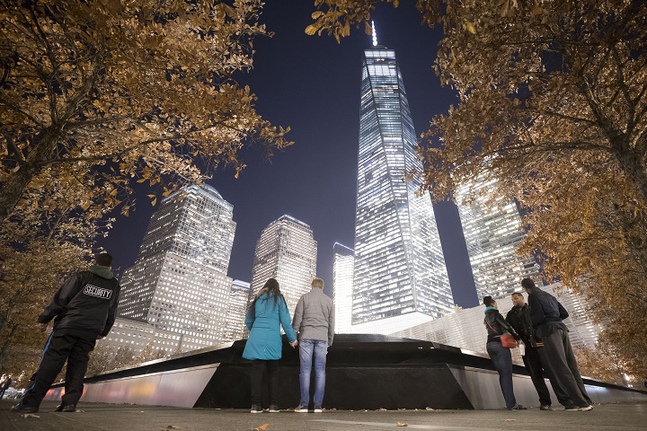 Groups of people stand around the 9/11 Memorial pool at night, beneath trees with rust-colored autumn leaves. One World Trade stands illuminated in the distance.