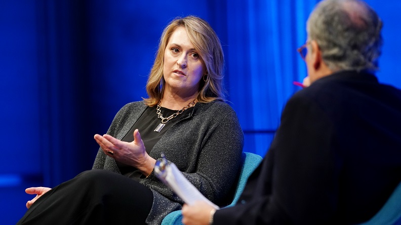 A woman wearing a gray blazer speaks to a man on a blue stage during a public program.