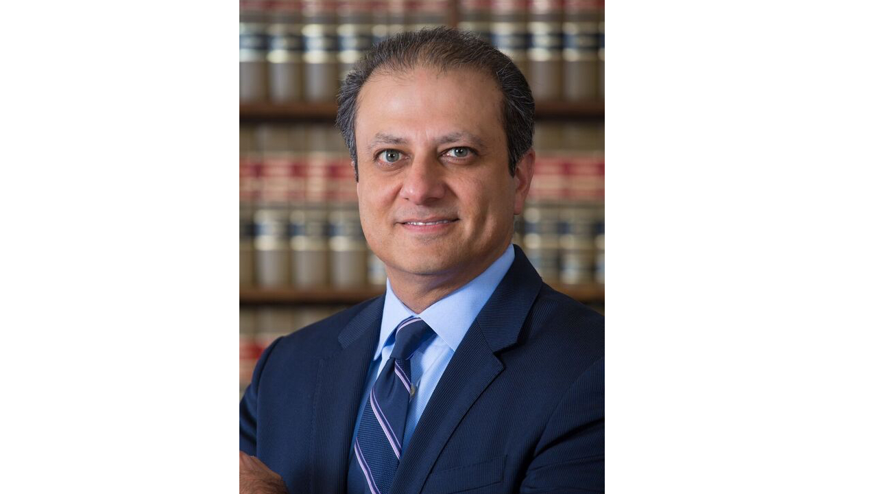 A headshot of Preet Bharara wearing a navy blue suit, standing in front of a wall of books.