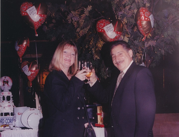 A woman with blonde hair and a man with a mustache toast with champagne in front of a white-tablecloth-covered table and a tree with heart-shaped balloons attached to it.