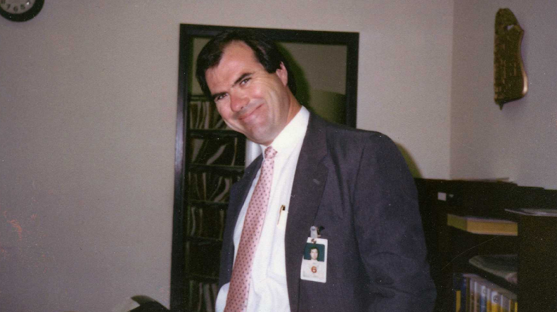 A man wearing a suit and tie and a professional badge grins with his head tilted to the side in an office.