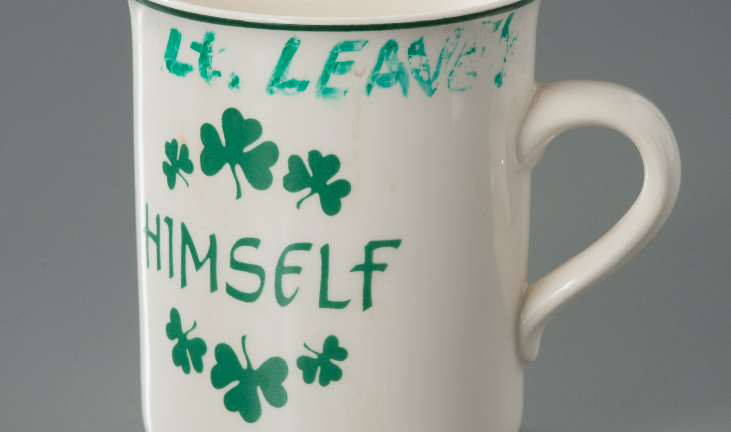 A mug belonging to FDNY Lieutenant Joseph Leavey is displayed on a gray surface at the museum. The white mug is adorned with green shamrocks. The word "himself" is printed in green on the mug. Leavey's name has been written in green marker on the lip of the cup.
