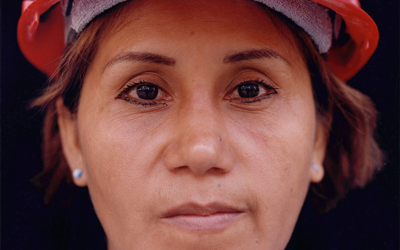 This close-cropped photograph shows a woman in a red hardhat, staring directly into the camera.