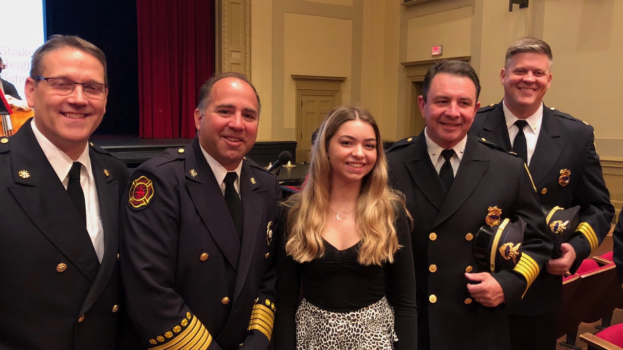 A blonde high schooler stands in between a group of four first responders in dress uniforms in a high school auditorium.