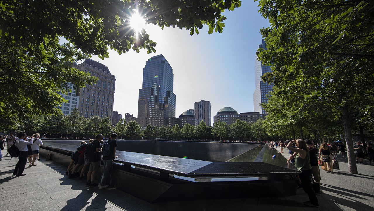 People mill about the 9/11 Memorial plaza on a summer's day. The sun shines through the trees.
