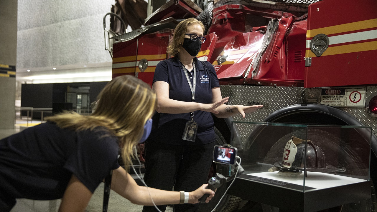 A woman wearing a face mask stands in front of the Ladder 3 firetruck in the Memorial Museum. Another woman films the scene with her smartphone.