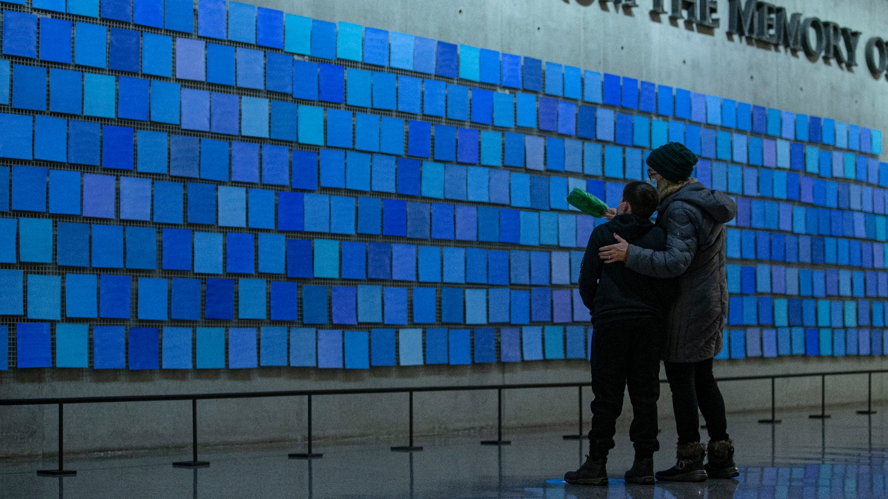 An older woman wraps her arm around a young boy's shoulder as they look at the inscription "No Day Shall Erase You From the Memory of Time.”  This quote from Book IX of "The Aeneid" by the Roman poet Virgil are are part of a larger art installation by artist Spencer Finch, titled “Trying to Remember the Color of the Sky on That September Morning.” The words appear amid a sea of blue tiles.