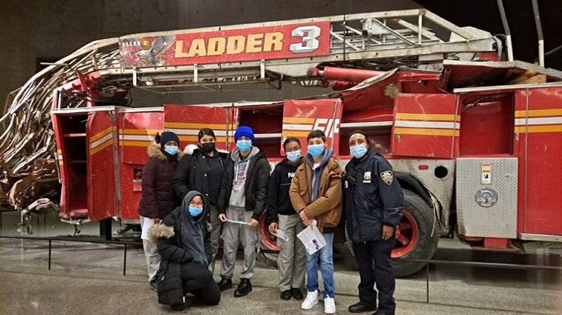 A group of young people wearing masks pose alongside an NYPD officer in front of the Ladder 3 firetruck in the 9/11 Memorial Museum.