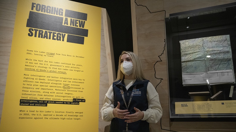 A female tour guide wears a mask and stands in front of wall text and a topographic artifact in the "Revealed: The Hunt for Bin Laden" exhibition.