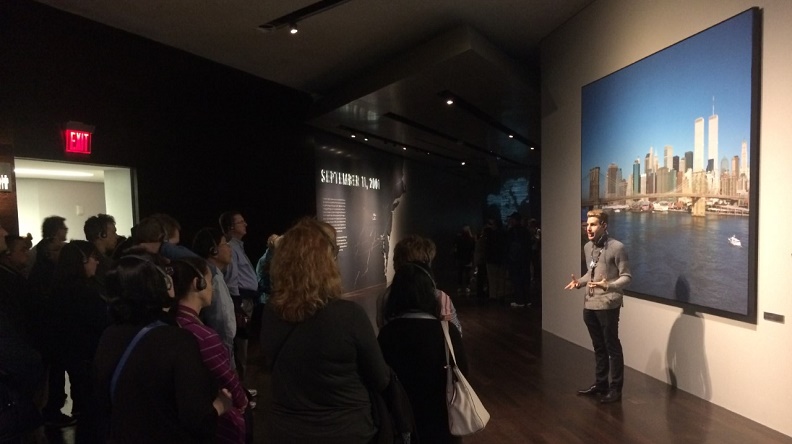 In this photograph, an interpretive museum guide stands before a blown-up photograph of the New York skyline pre-9/11, giving a tour to a group of museum visitors, who listen through headsets.