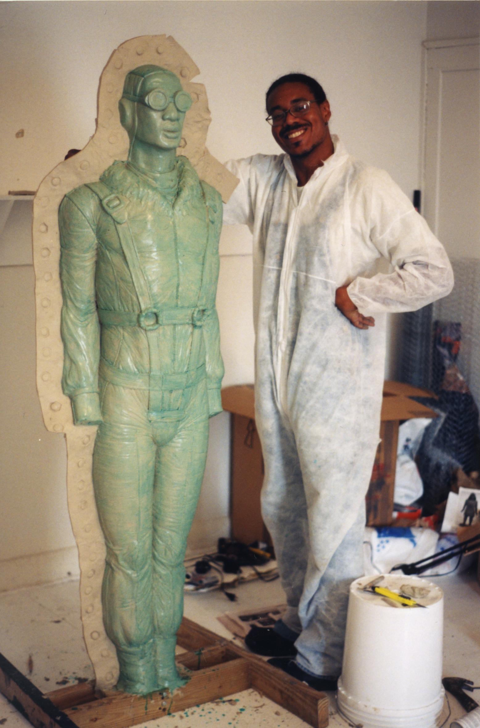 A smiling artist dressed in white stands to the right of a green sculpture.