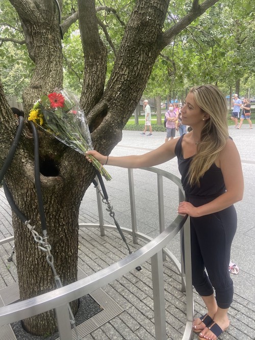 A young woman with blonde hair, dressed in black, places a colorful bouquet at the Survivor Tree on the Memorial.