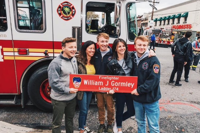 A smiling family - three sons, a daughter, and a mother - hold up a red sign with the name WILLIAM J GORMLEY on it in white letter