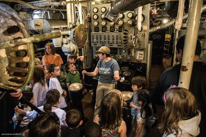 A woman with short hair and a khaki baseball cap shows a group of small children around a ship's engine room