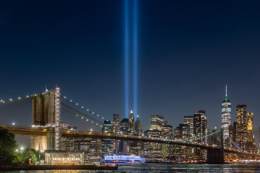 The Tribute in Light, blue beams representing the fallen Twin Towers, standing above the Brooklyn Bridge and Manhattan skyline at night