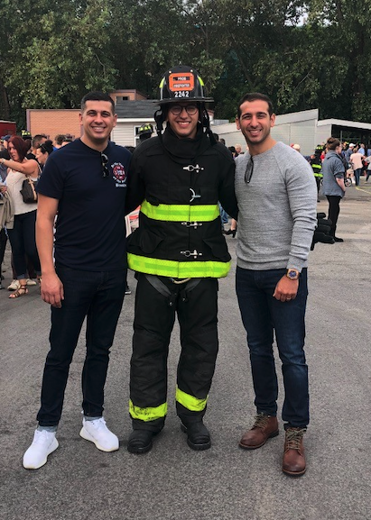 Three young men stand with their arms around each other; at center is a uniformed FDNY firefighter. At left, a young man wearing a dark t-shirt and dark jeans. At right, a young man wearing a gray sweater and jeans.