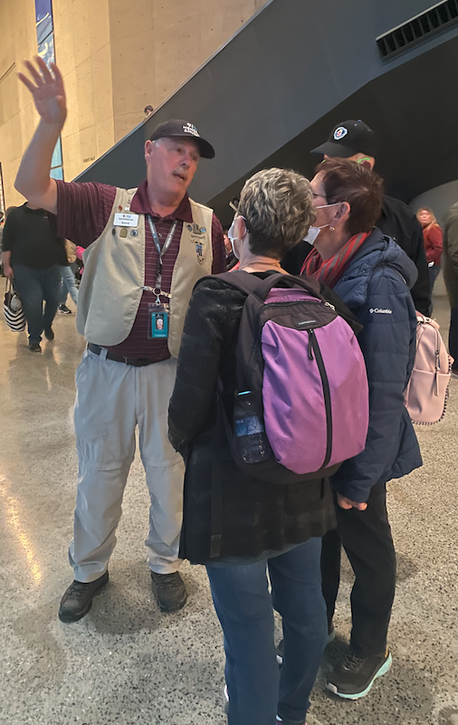 A gray-haired man with a mustache, wearing a burgundy top and khaki volunteer vest, guides some female visitors wearing backpacks.