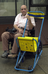 A man in a white shirt and khaki pants sits to the left of a portable wheelchair with blue finish and yellow seat. 