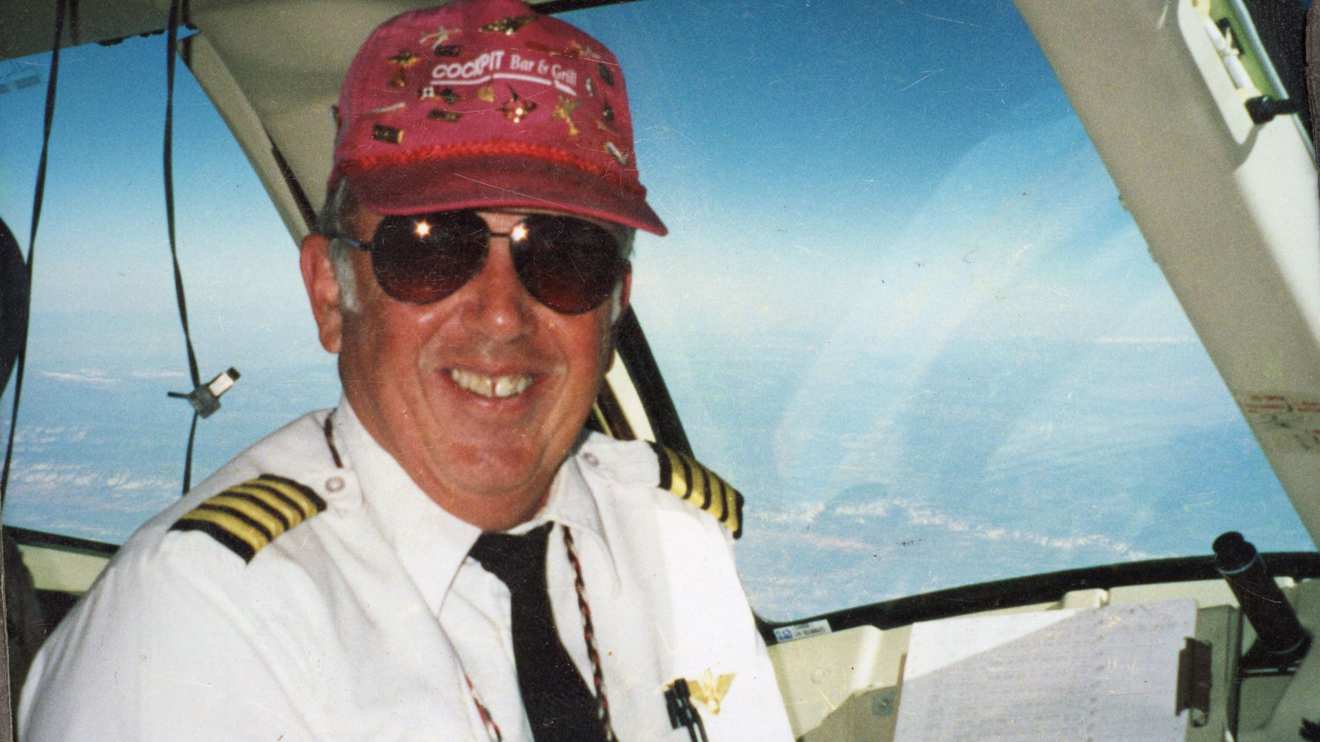 A smiling man in a cockpit, wearing a pilot's uniform and a white baseball cap