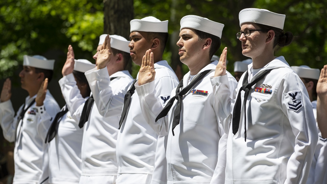 Formally dressed members of the U.S. Navy hold up their right hands as they stand at Memorial plaza.