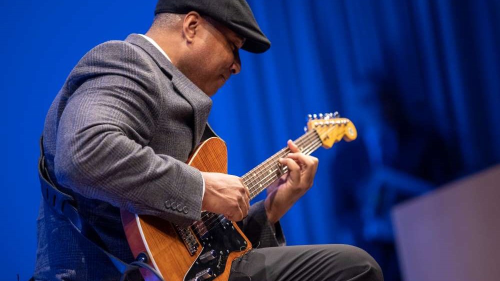 Former Yankees player Bernie Williams plays an electric guitar on stage in the Auditorium as part of a public program. He is wearing a suit, tie, and hat.