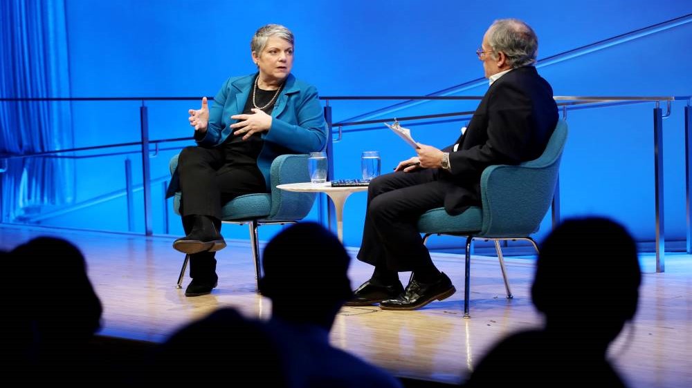 Former Secretary of the Department of Homeland Security Janet Napolitano speaks onstage as she gestures with both hands. Clifford Chanin, the executive vice president and deputy director for museum programs, listens as he sits next to her with a clipboard. The silhouettes of several audience members are visible in the foreground.