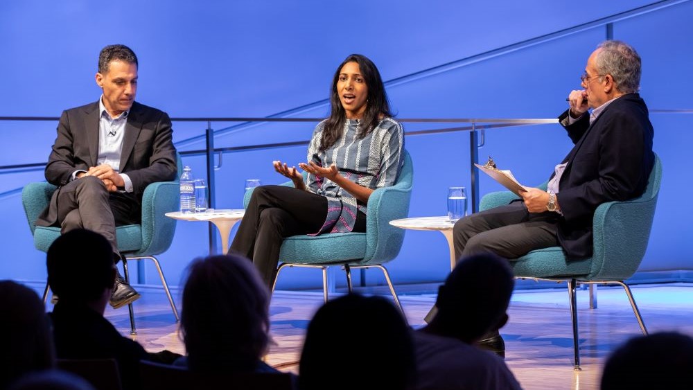 Vidhya Ramalingam, founder of Moonshot CVE, holds out her hands as she speaks onstage at the Museum Auditorium. Hany Farid, Dartmouth computer science professor and senior advisor to the Counter Extremism Project, listens with his hands in his lap to her left. Clifford Chanin, the executive vice president and deputy director for museum programs, sits to her right holding a clipboard and listening on. Audience members in the foreground are silhouetted by the white and blue lights onstage.