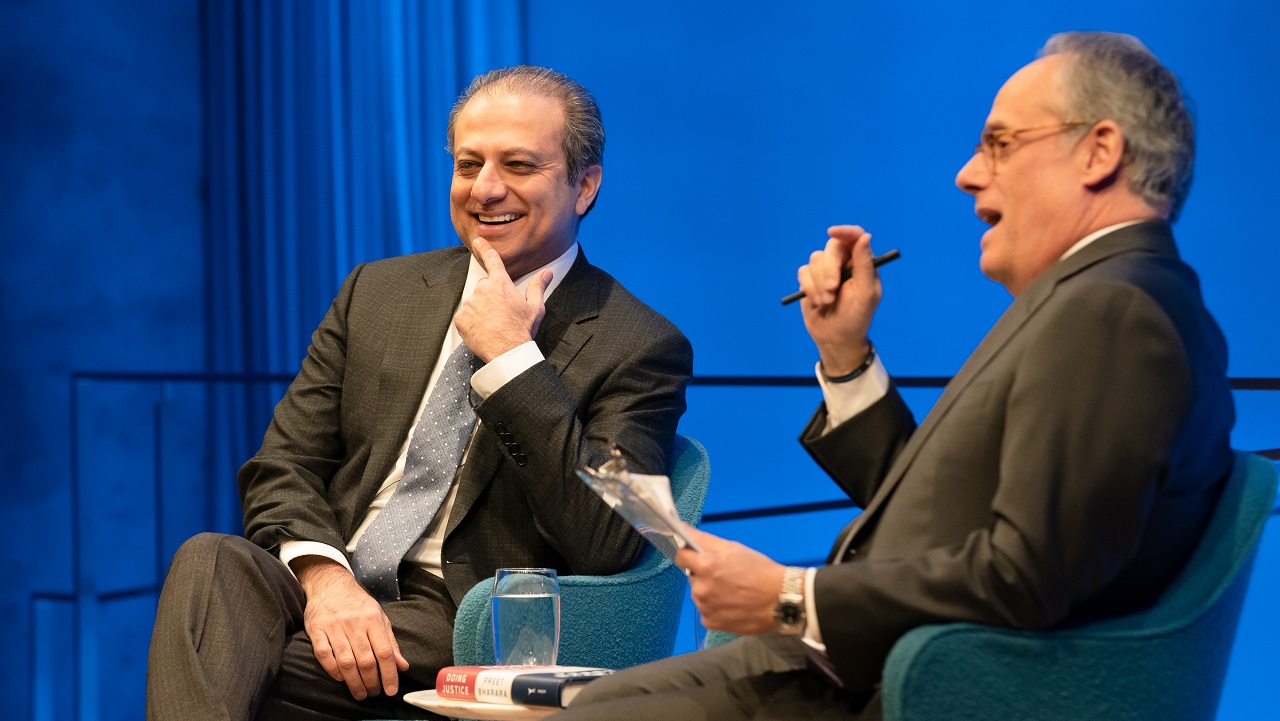 Preet Bharara, the former U.S. attorney for the Southern District of New York, smiles as he sits on stage with moderator Clifford Chanin. Bharara is holding his left index finger to his face as he listens to Chanin speak. Chanin is holding a clipboard in his left hand and a pen in his right hand. The two men are seated in front of a wall that is lit blue from the stage lights.
