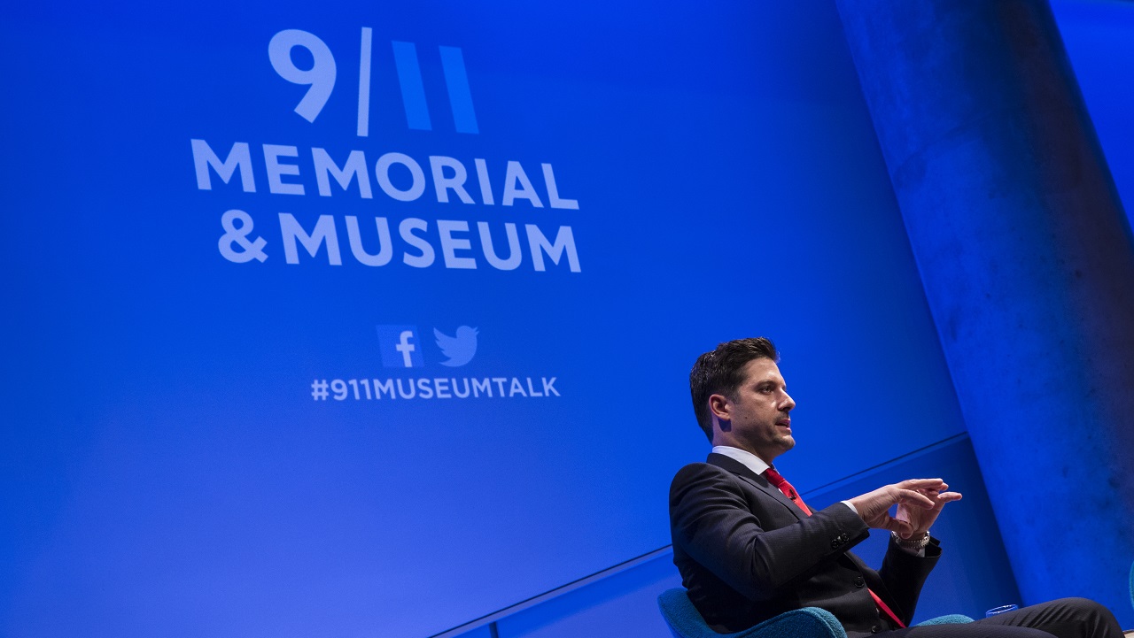 In this photograph, a man in a suit and red tie sits on a blue-lit auditorium stage, with “9/11 Memorial & Museum #911museumtalk” projected onto the wall behind him. He is shown in profile, gesturing with his hands to an unseen audience.