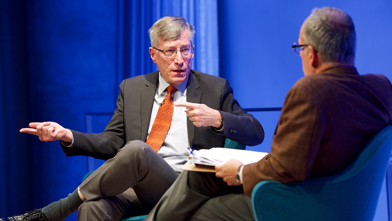 In this close-cropped photograph, two men sit on an auditorium stage. One man in a suit and orange tie gestures by pointing both index fingers to the right while the other man, whose back is to the camera, listens.