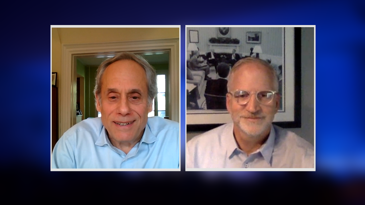 A screenshot from a video conference call shows two men with graying hair. A man to the left of the frame is listening intently with his hands clasped in front of him. To the right, a man in glasses is speaking.