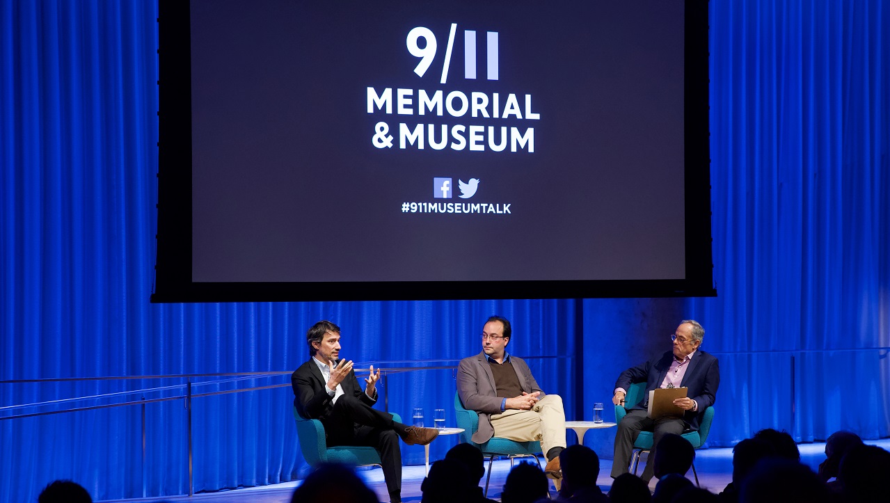 Three men in suits sit on a blue-lit auditorium stage. Behind them "9/11 Memorial & Museum #museumtalk" is projected on a screen behind them.
