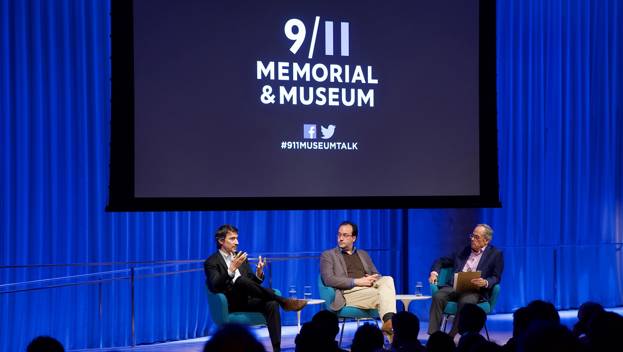 Three men in suits sit on a blue-lit auditorium stage. Behind them "9/11 Memorial & Museum #museumtalk" is projected on a screen behind them.