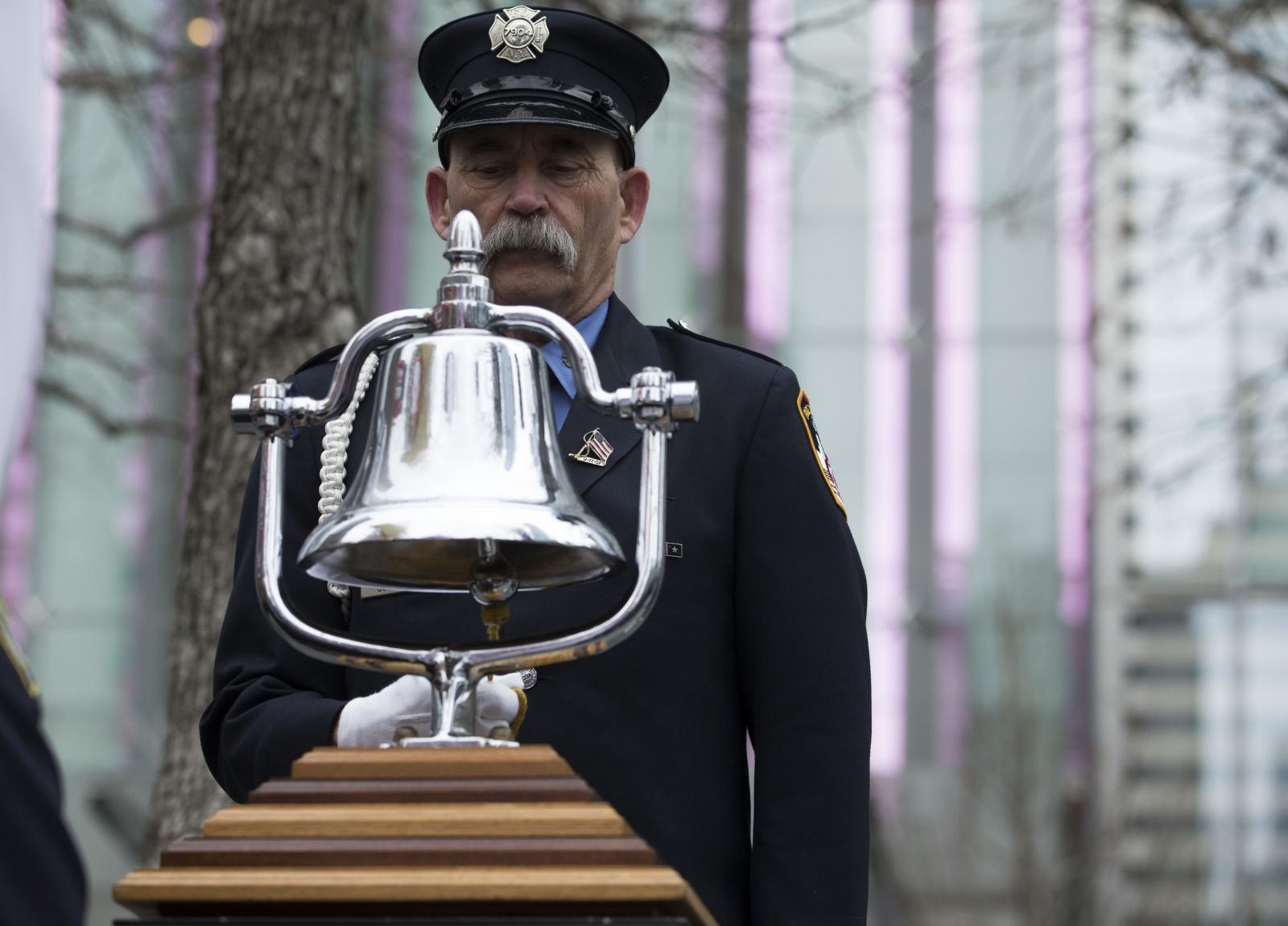 Firefighter with mustache, in formal uniform, stands in front of a silver bell