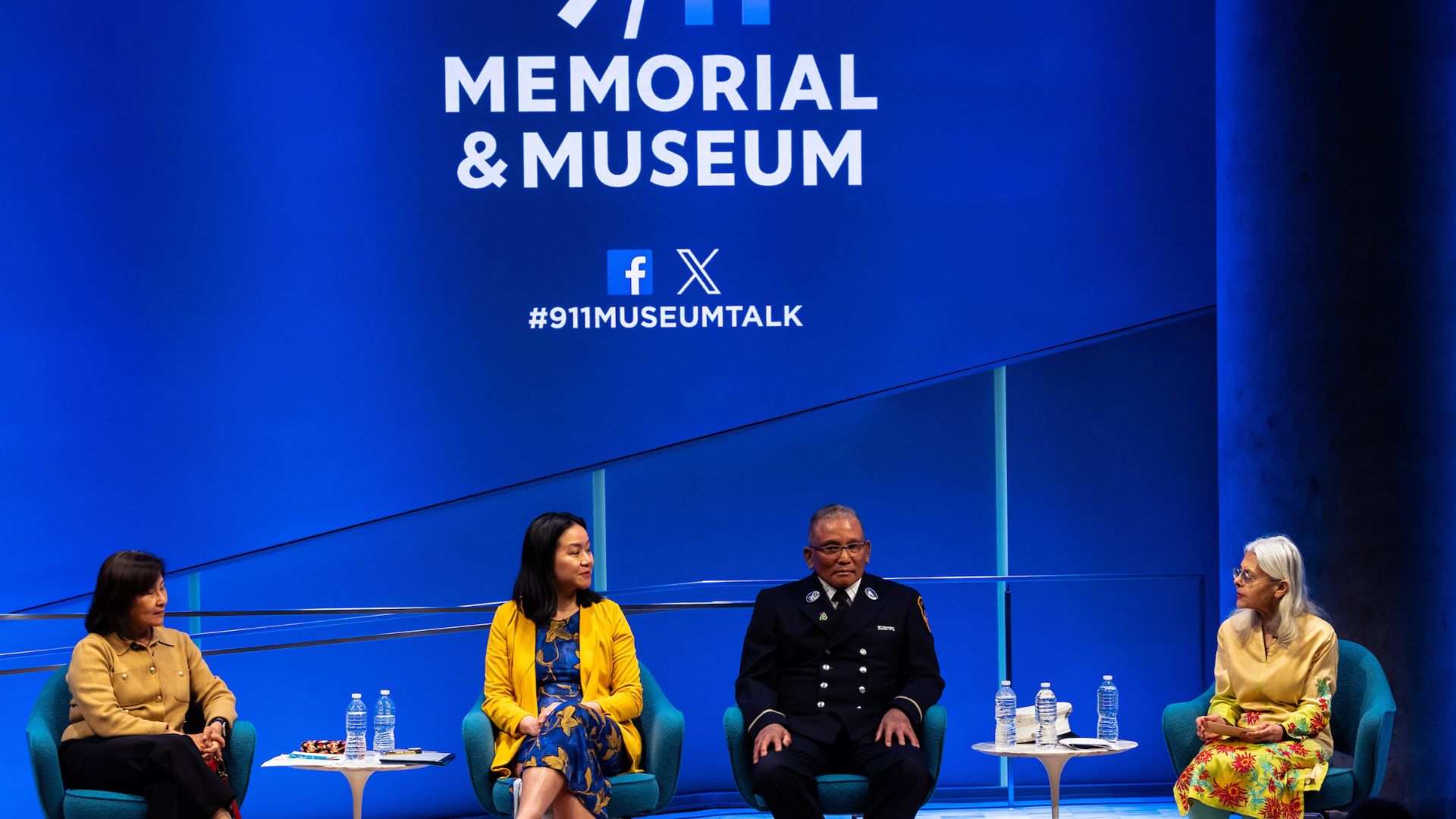 Four panelists seated on stage in front of a bright blue backdrop with the 9/11 Memorial & Museum logo visible. 