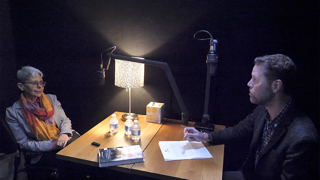At a table in a dimly lit room, a woman with her hands on her lap sits across from a man with a pencil and paper. There is a lamp on the table and microphones are positioned in front of the man and woman.