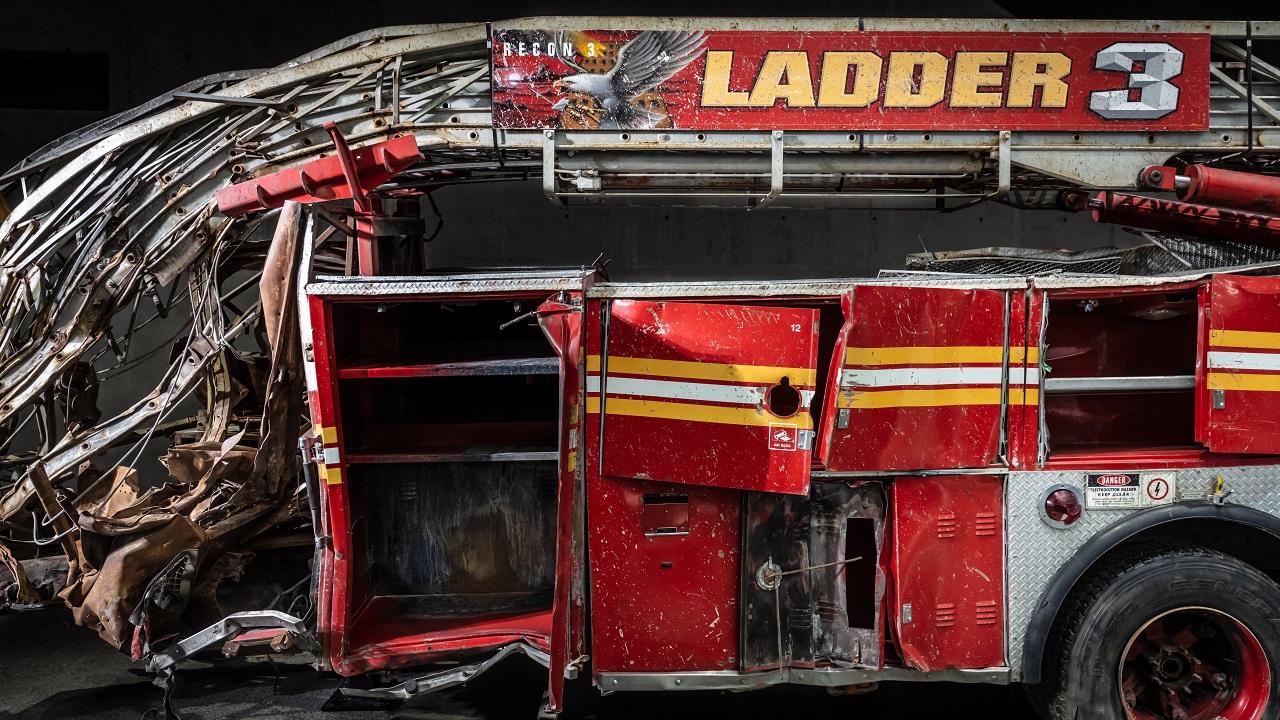 The heavily damaged firetruck of Ladder Company 3 sits in the Museum. This close-up view shows the bright red vehicle’s twisted ladder and broken compartment doors.