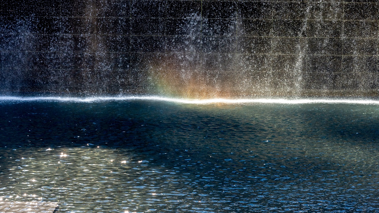 A rainbow forms on the surface of one of the memorial pools as water cascades down in the background.