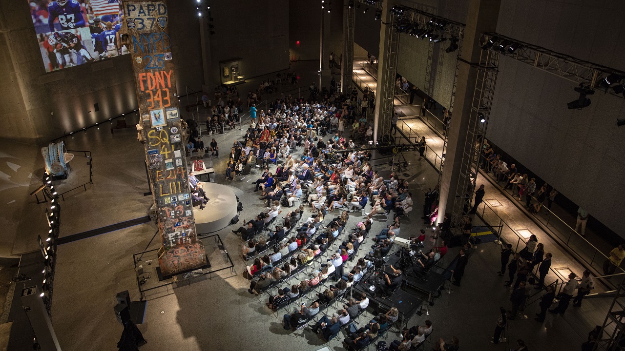 Dozens of seated people watch people speaking on a stage beside the Last Column at Foundation Hall. A video is projected on a wall and the lighting in the hall has been dimmed for the event.