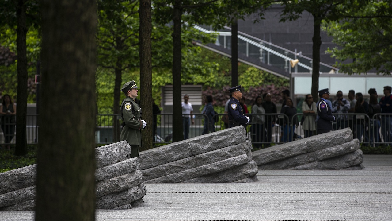 Three officials in formal uniforms stand beside three granite monoliths at the Memorial Glade. A crowd of people has gathered in the shade of trees behind them.