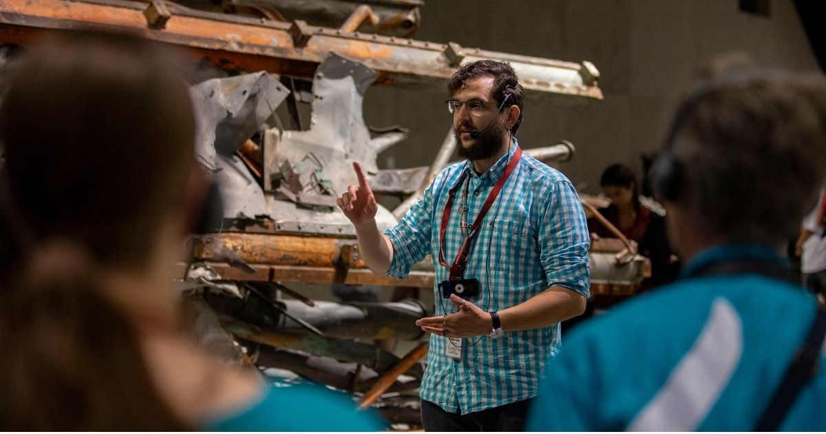 A bearded young male tour guide, standing in front of a mangled segment of an antenna recovered from the World Trade Center, gestures as talks to visitors on a tour of the 9/11 Memorial Museum.