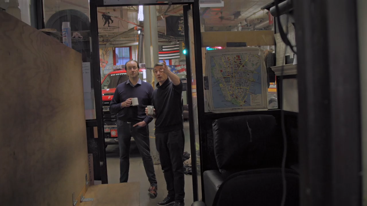 Two men standing in an inner doorway of a fire station, holding cups of coffee and talking. Behind them are a fire truck and an American flag with red firefighter stripe.