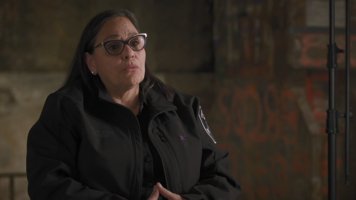 A woman wearing a black FDNY jacket with a white badge on the shoulder sits and talks.