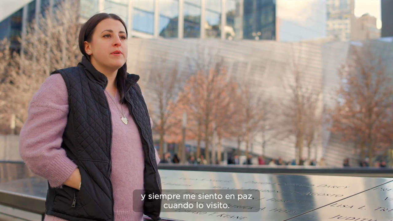 Christine Fiorelli Epstein stands near hear father's name inscribed on the 911 Memorial wearing a pink sweater, black vest, and silver heart necklace. Her long brown hair is tucked into her vest and her hands are in her pockets. 