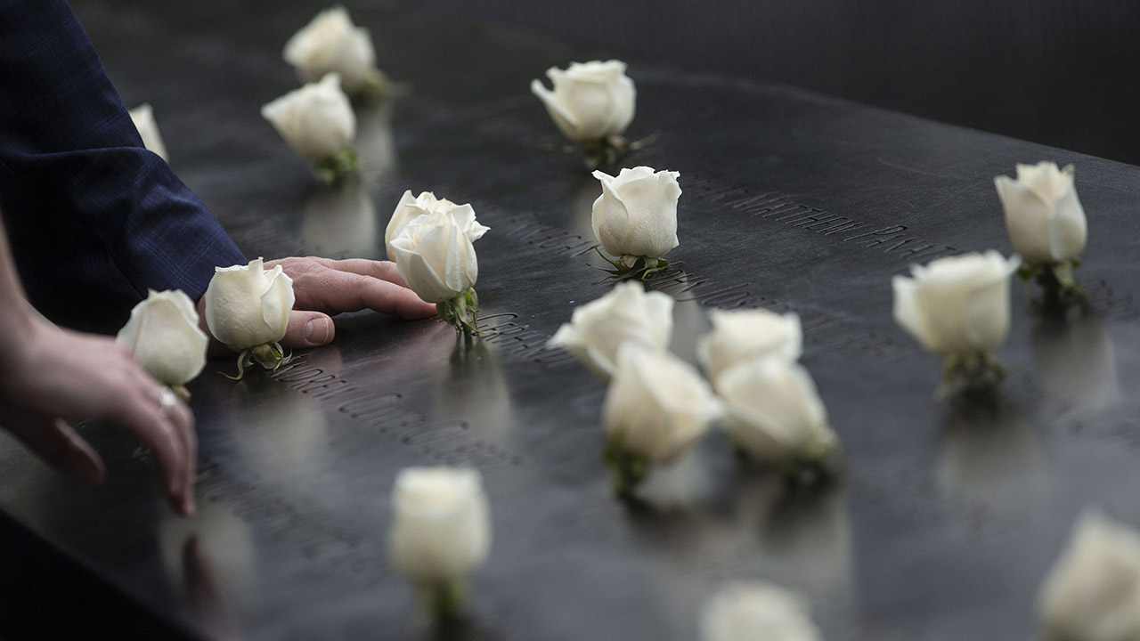 Two people have each placed a hand on a bronze parapet at the Memorial. They are next to about a dozen white roses that have been placed at the names of victims.