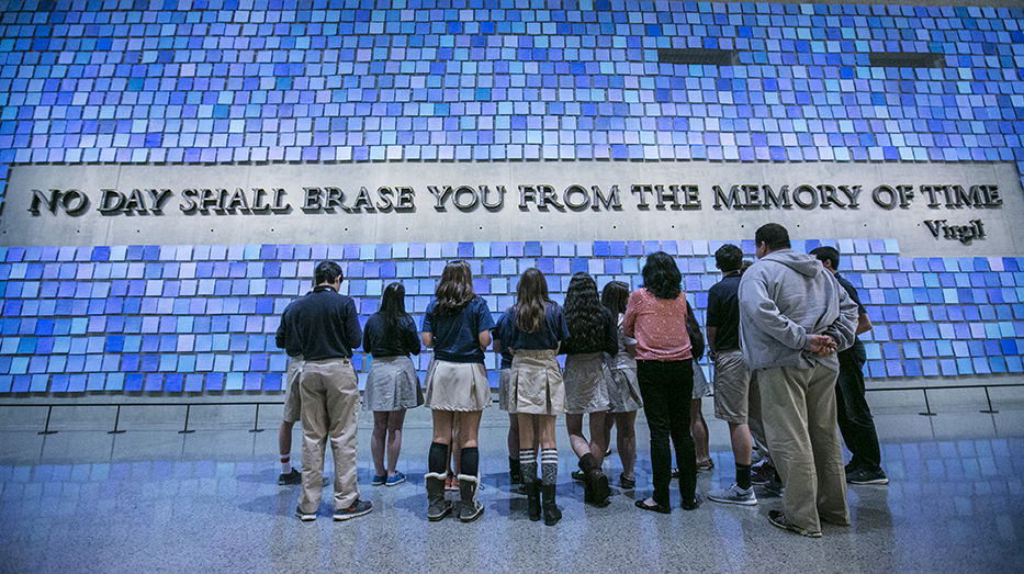 A group of students in school uniforms looks up at a large inscription on the wall that reads “No day shall erase you from the memory of time.” The quote from Virgil’s epic poem The Aeneid is surrounded by 2,983 unique blue tiles that comprise "Trying to Remember the Color of the Sky on That September Morning" by Spencer Finch. 