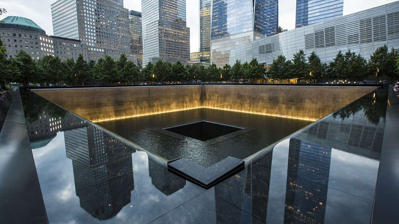 The 9/11 Memorial reflects buildings and clouds as the sun sets over the South Pool of Memorial Plaza.