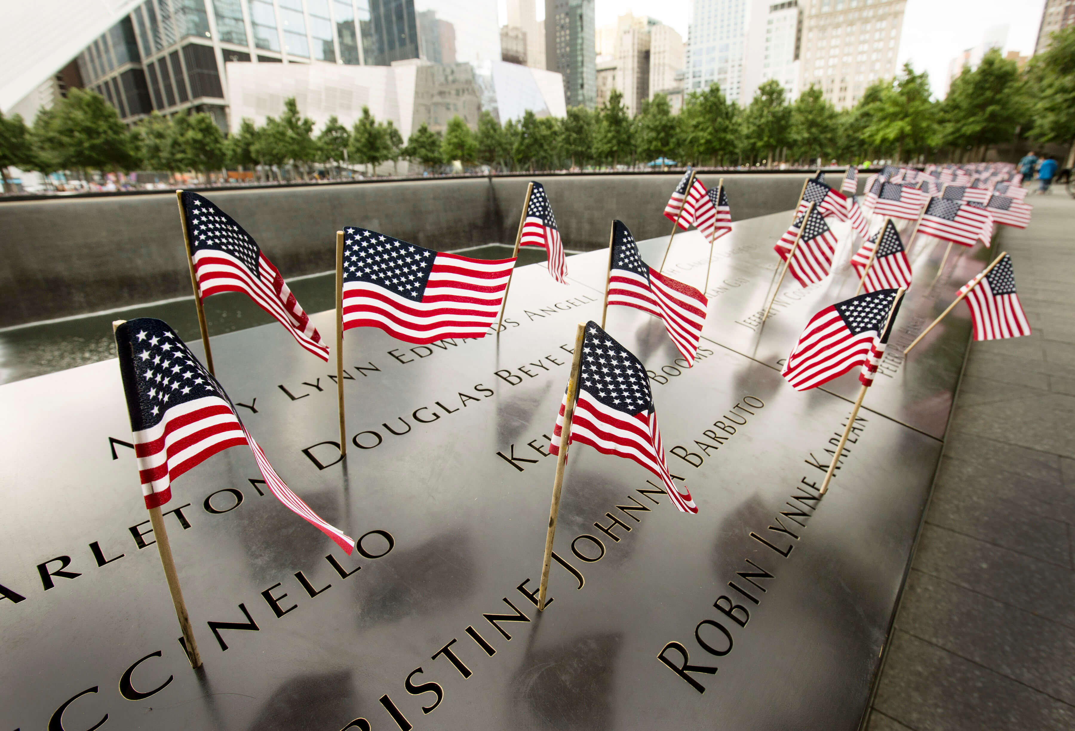 Dozens of American flags wave at the names on the bronze parapets of the Memorial. Buildings and trees are in the background.