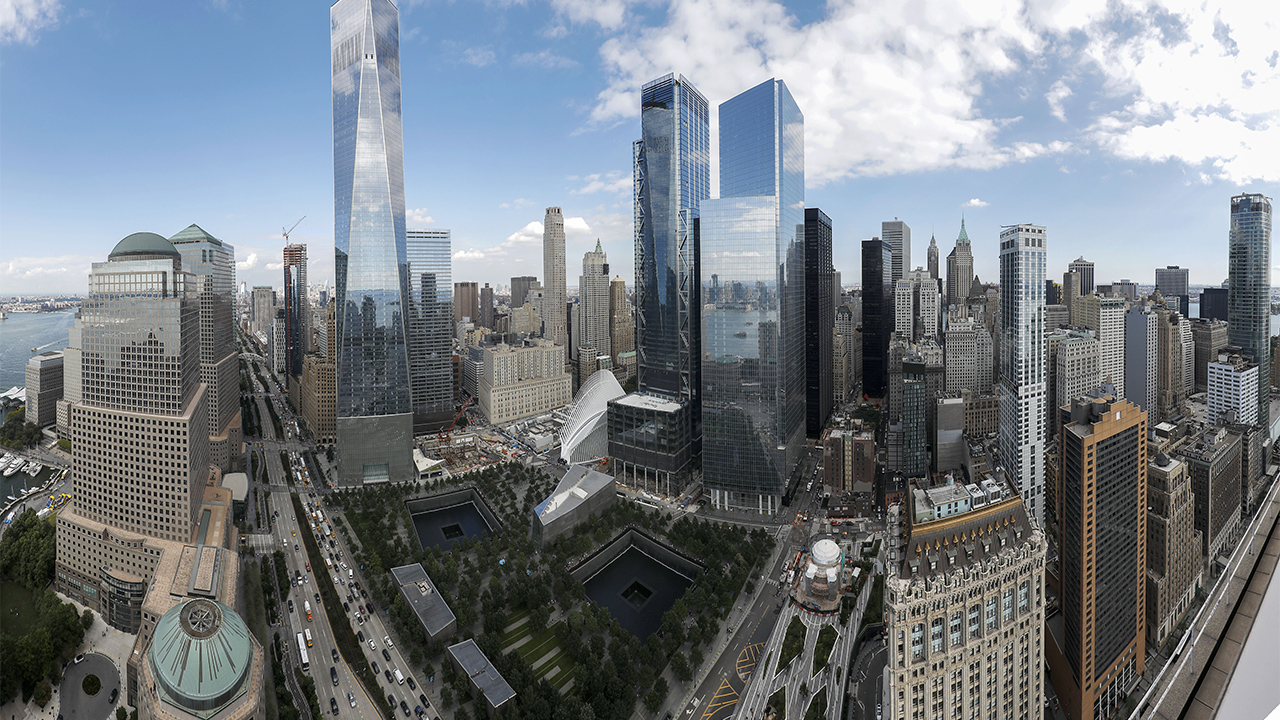 An aerial view over World Trade Center on a partly cloudy day. One World Trade Center towers over the Memorial with the reflecting pools at its center. The skyline of lower Manhattan stands to the right.